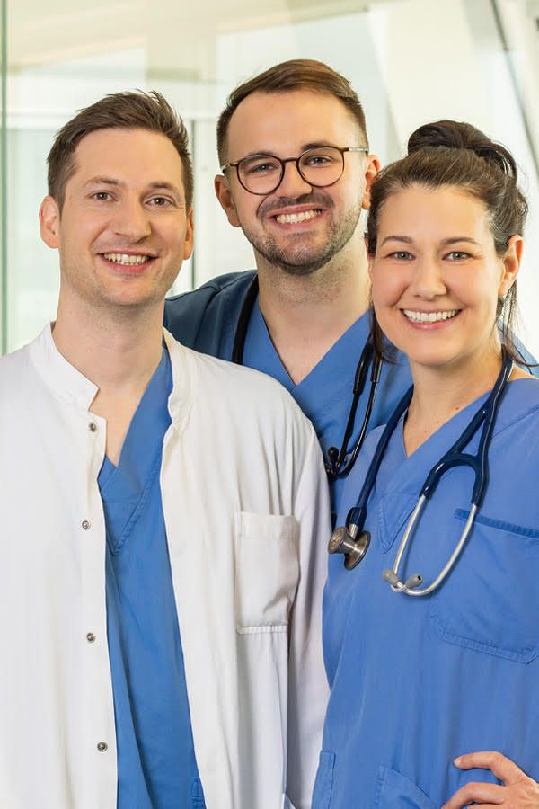 Drei medizinische Fachkräfte in OP- und Arztkleidung stehen lachend in einem hellen Krankenhausgang. Ein Mann in grünem Kittel steht in der Mitte, die anderen beiden flankieren ihn links und rechts.