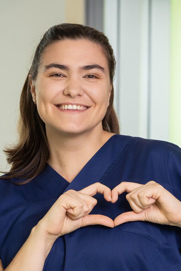 Eine Pflegerin oder medizinische Fachkraft in dunkelblauer Kleidung formt mit den Händen ein Herzsymbol und lächelt freundlich in die Kamera. Der Hintergrund zeigt eine helle Krankenhausumgebung.