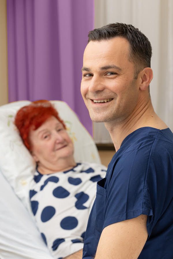 Ein Pfleger in dunkelblauer Kleidung sitzt lächelnd neben einem älteren weiblichen Patienten im Krankenhausbett. Die Patientin mit roten Haaren trägt ein weißes Hemd mit blauen Punkten.