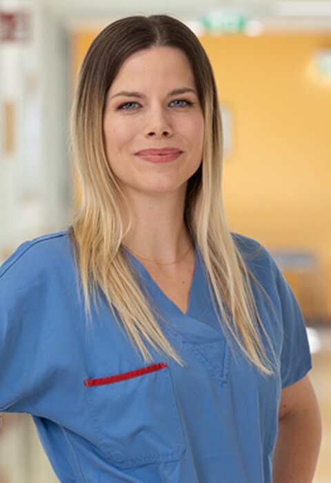 Eine junge medizinische Fachkraft mit langen, offenen, blond-braunen Haaren trägt blaue OP-Kleidung und steht selbstbewusst in einem hellen Krankenhausflur, während sie leicht lächelt.